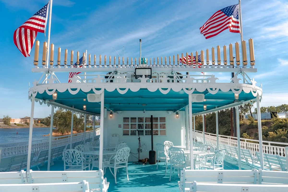 The open top deck of a riverboat with white wrought-iron chairs under a turquoise canopy and American flags